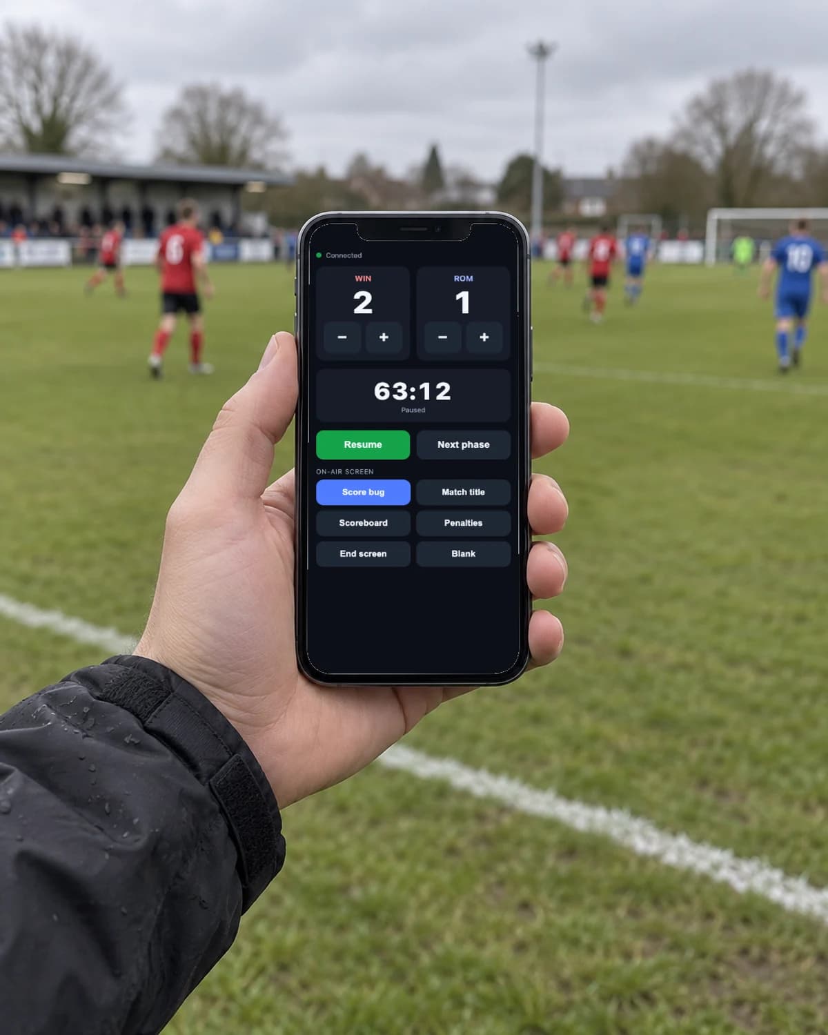 A hand holding a phone at the touchline, showing the PlayOverlay phone remote with the score and clock