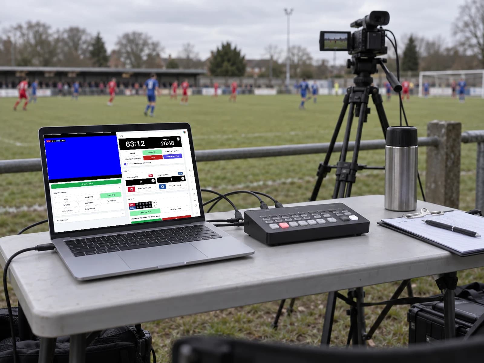 A laptop running PlayOverlay beside a compact HDMI video switcher, with a camera on a tripod beside the pitch